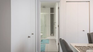 Un couloir mène à une salle de bain dotée d’une douche en verre, d’une porte blanche et d’un coin repas avec une table et des chaises.