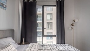 Une chambre à coucher avec un lit, une fenêtre avec des rideaux, une vue sur un immeuble et un luminaire fixé au mur.