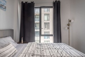 A bedroom with a bed, a window with curtains, a view of a building, and a wall-mounted light fixture.