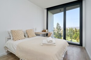 Un dormitorio con cama, almohadas, una mesita de noche y vistas a través de un ventanal grande.