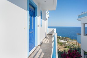 Un balcone stretto con una porta blu, affacciato su una zona costiera con cielo azzurro e mare limpido.