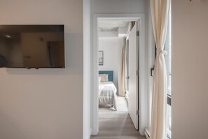 Un couloir mène à une chambre à coucher équipée d’un lit, de rideaux et d’un téléviseur fixé au mur.