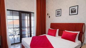 Hotelkamer met een bed, rode gordijnen, een balkon met een tafel en stoelen, en twee ingelijste foto’s aan de muur.