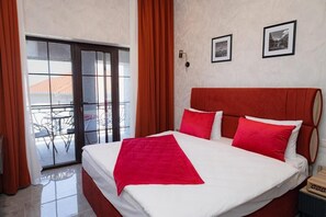 Habitación de hotel con una cama, cortinas rojas, un balcón con mesa y sillas, y dos cuadros en la pared.