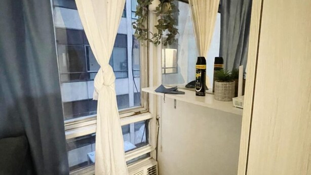 Une petite pièce, de taille modeste, avec une fenêtre, un radiateur, un miroir et une étagère sur laquelle se trouvent une plante et une bouteille.