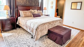 A bedroom with a large bed, a nightstand, a bedside table, a footstool, a patterned rug, a ceiling fan, and a door leading to another room.