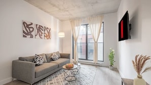 Una sala de estar moderna con un sofá gris, arte abstracto en la pared, una mesita redonda con un frutero, una planta en maceta y un televisor montado.