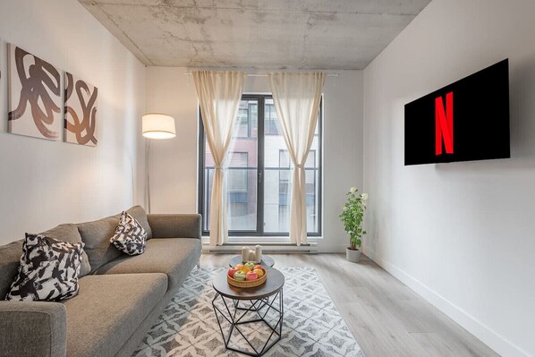 Uma sala de estar moderna com um sofá, uma mesa de centro com frutas, uma planta em vaso e uma TV fixada na parede.