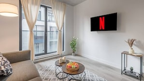 Una sala de estar moderna con un amplio ventanal, un sofá, una mesa de centro con fruta, un televisor montado en la pared mostrando el logotipo de Netflix y una pequeña planta.