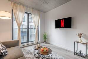 A modern living room with a large window, a sofa, a coffee table with fruit, a wall-mounted TV displaying the Netflix logo, and a small plant. - 1 bedroom old port- F1 Weekend (Montreal)