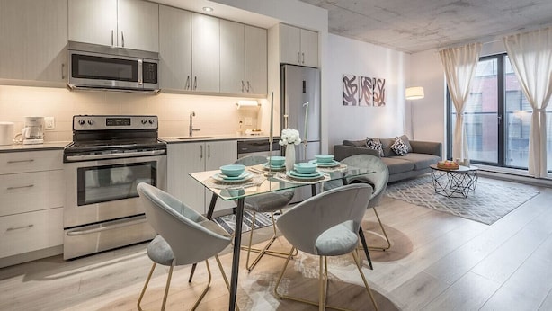 Una cocina moderna con electrodomésticos de acero inoxidable, un comedor con mesa de vidrio y sillas grises, y una sala de estar con sofá y un ventanal grande.