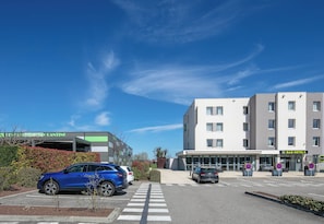 A modern building with a parking lot, a blue car, and a B&B hotel sign. - B&B Hotel Lyon Aeroport Saint Quentin Fallavier (Saint-Quentin-Fallavier)