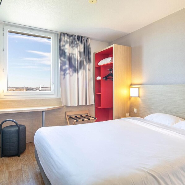 A hotel room with a bed, a window with curtains, a red storage unit, a bedside table, and a wall-mounted lamp. - B&B Hotel Lyon Aeroport Saint Quentin Fallavier (Saint-Quentin-Fallavier)
