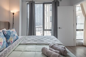 A bedroom with a bed, pillows, a floor lamp, a door, and a view of buildings through a window. - QUaint 2 BR- Parking -Square Vic (Montreal)