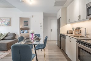 A modern kitchen with stainless steel appliances, a dining area with a glass table, and a living space with a sofa and a painting. - QUaint 2 BR- Parking -Square Vic (Montreal)
