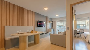 Ein modernes Wohnzimmer mit einem Holzesstisch, einer Couch, einem an der Wand montierten Fernseher und Blick ins Freie durch große Fenster.