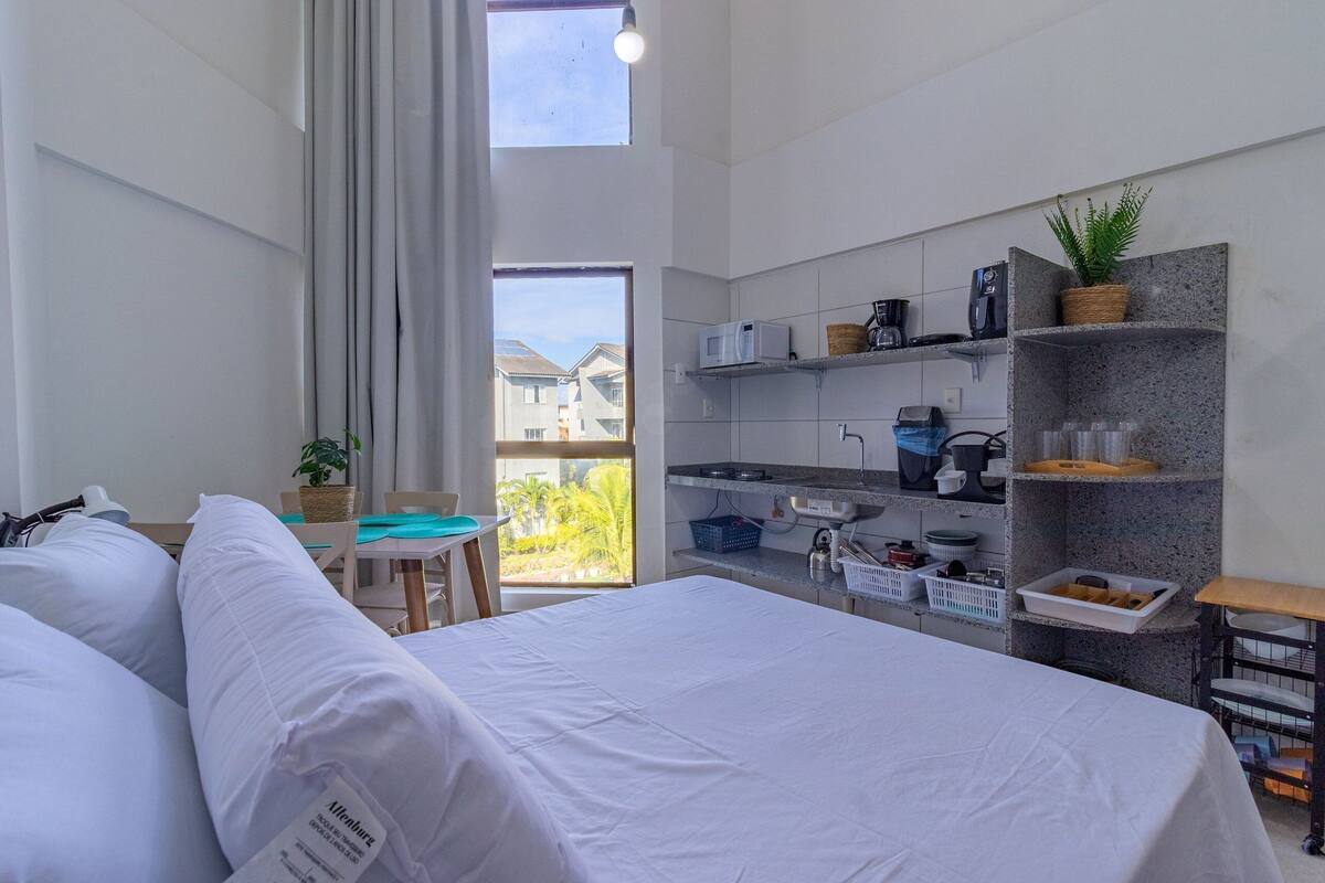 Una habitación de hotel moderna con una cama grande, una pequeña mesa de comedor, una kitchenette y un balcón con vistas a casas.