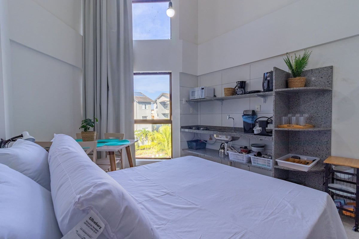 A modern hotel room with a large bed, a small desk, a kitchenette, and a balcony with a view.