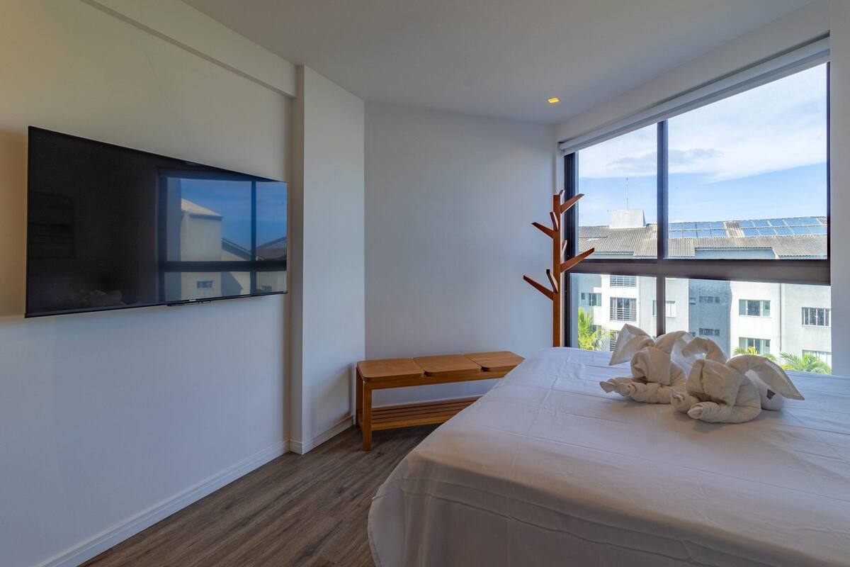 Una habitación de hotel moderna con una cama grande, un televisor de pantalla plana montado en la pared, un banco de madera y vistas a edificios a través de un ventanal amplio.