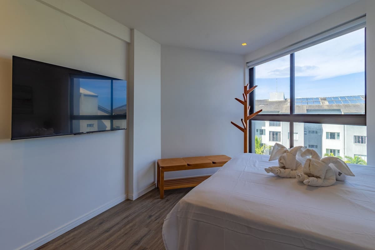 A modern hotel room with a large bed, a flat-screen TV mounted on the wall, a wooden bench, and a view of buildings through a large window.