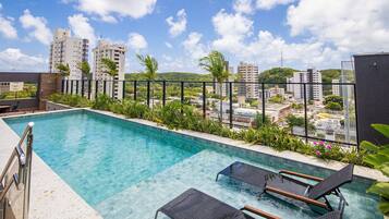 A rooftop pool with clear blue water, surrounded by greenery and overlooking a cityscape with tall buildings.