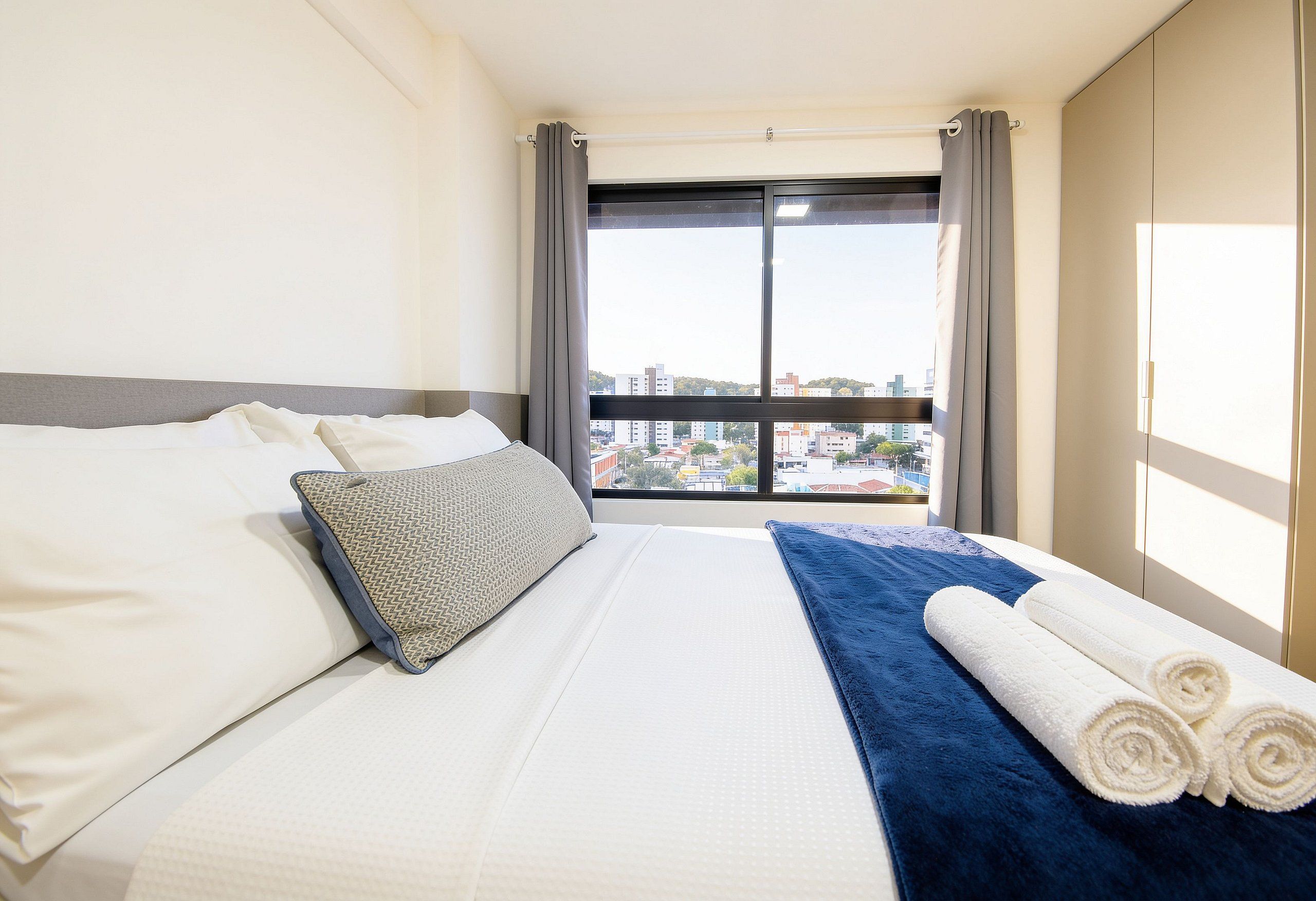 Quarto de hotel com cama, almofadas, toalha azul e vista para a paisagem urbana pela janela.