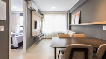 A modern, well-lit room with a dining table, chairs, a sofa, a TV, and a bed in the background.