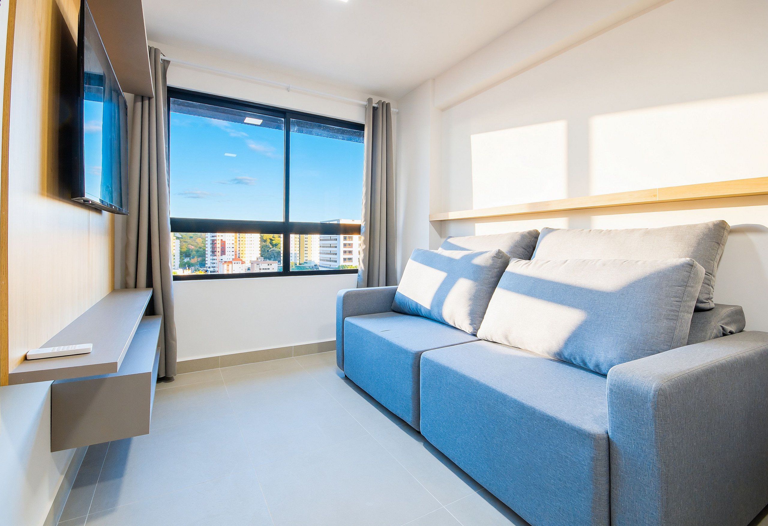 A modern living room with a grey sofa, a flat-screen TV mounted on the wall, and a window with a view of buildings.