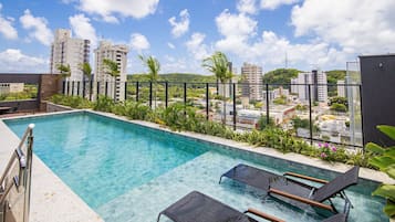 A rooftop pool with clear blue water, surrounded by greenery and overlooking a cityscape with tall buildings.