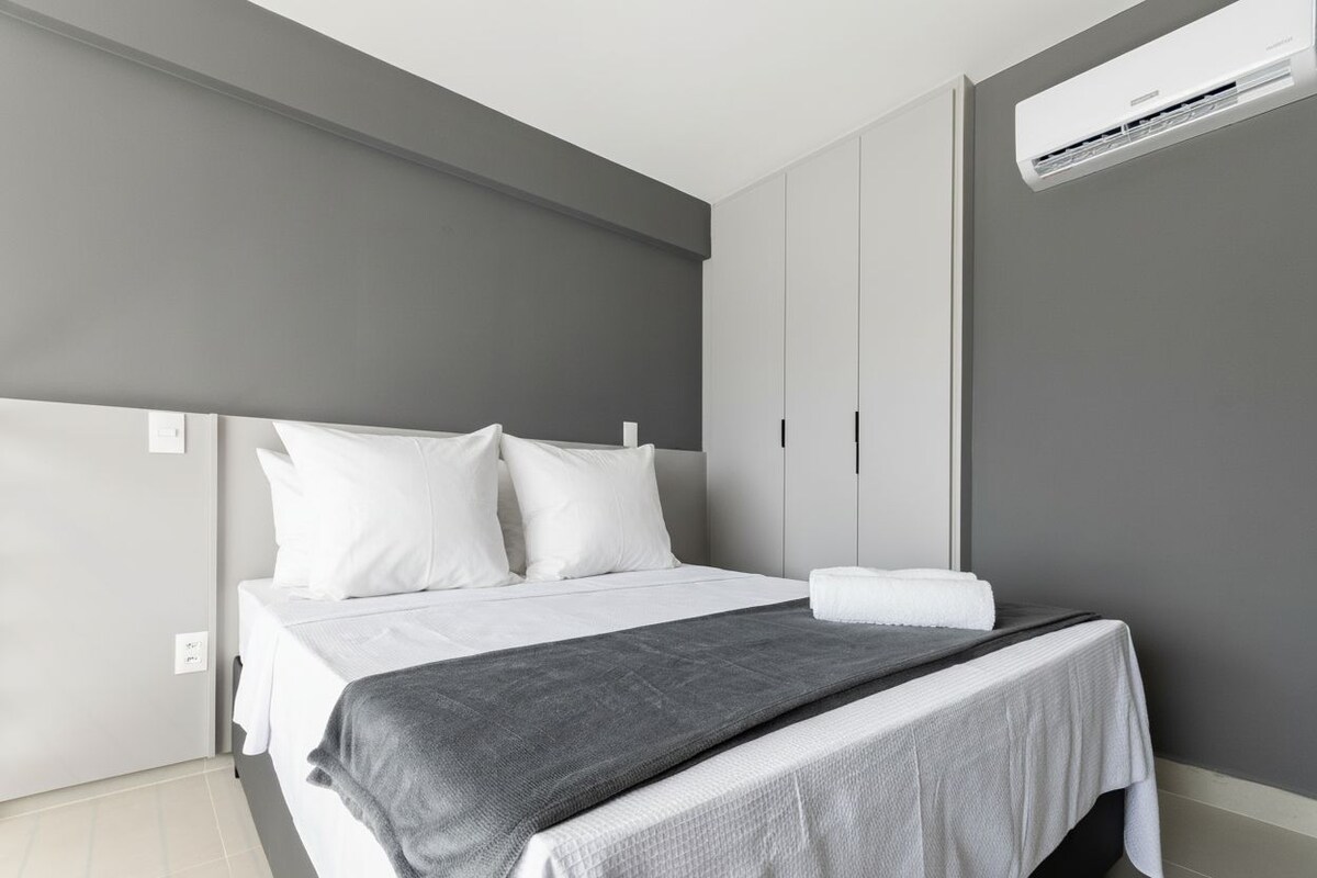 Une chambre d’hôtel moderne avec un lit, des murs gris, du linge de lit blanc et un climatiseur fixé au mur.