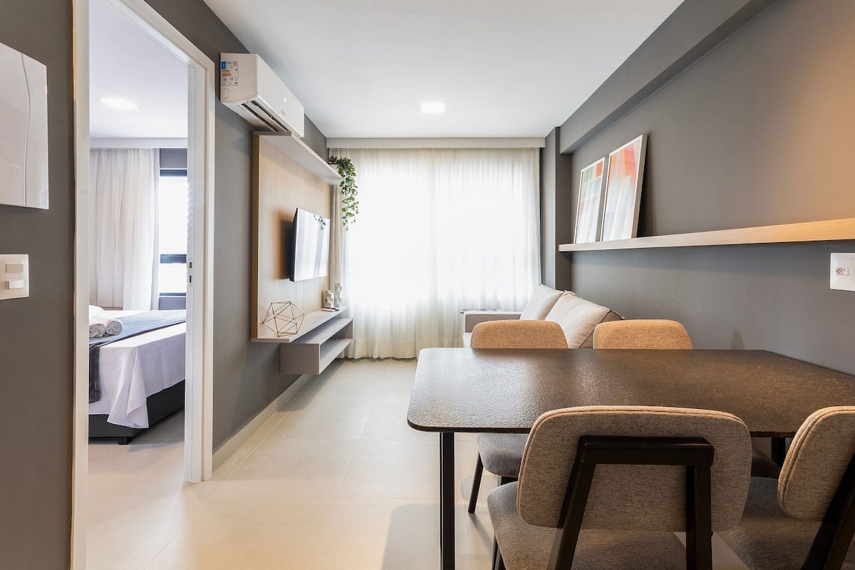 A modern, well-lit room with a dining table, chairs, a sofa, a TV, and a bed in the background.