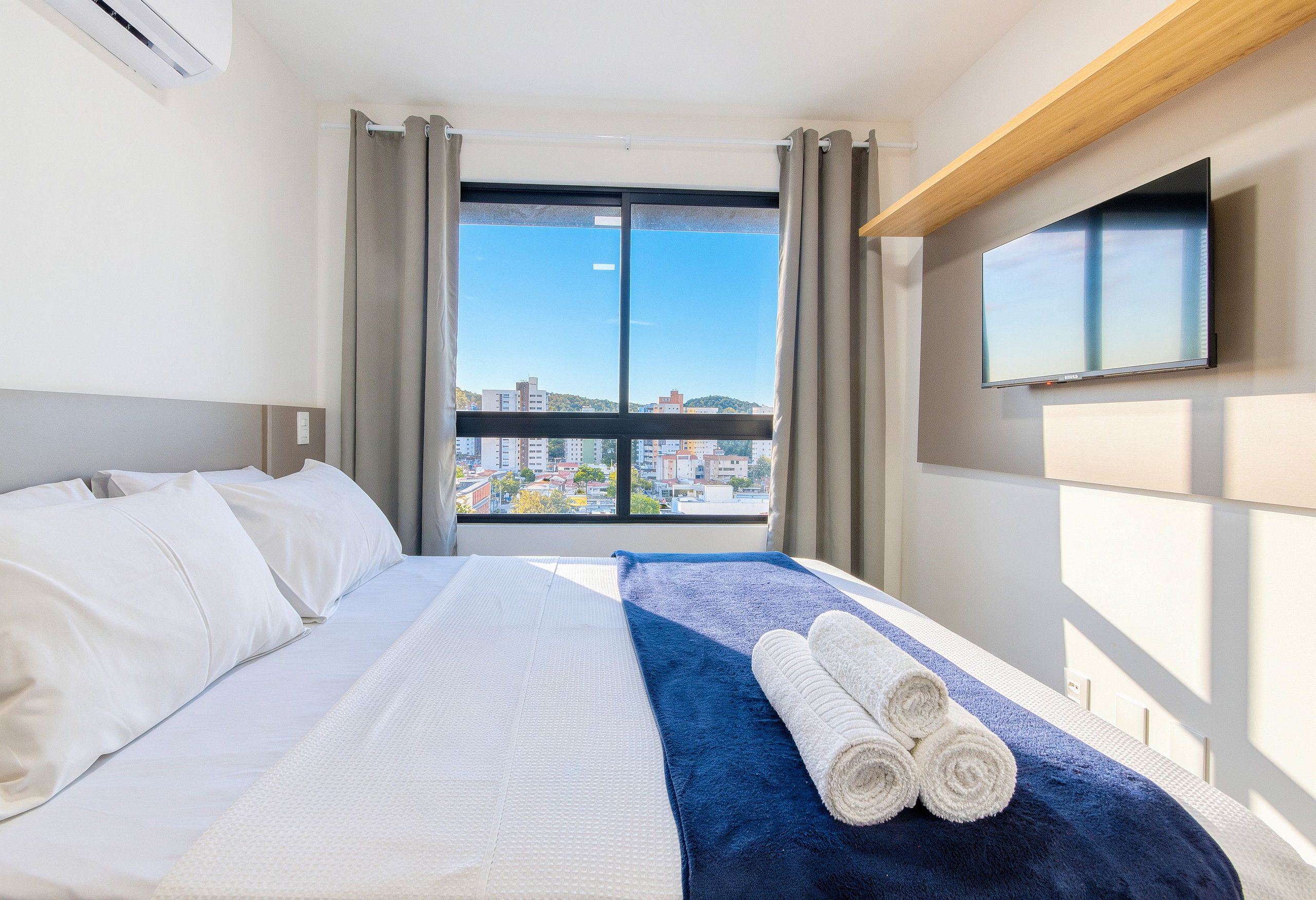 A hotel room with a bed, a window with curtains, a flat-screen TV mounted on the wall, and a view of the cityscape.