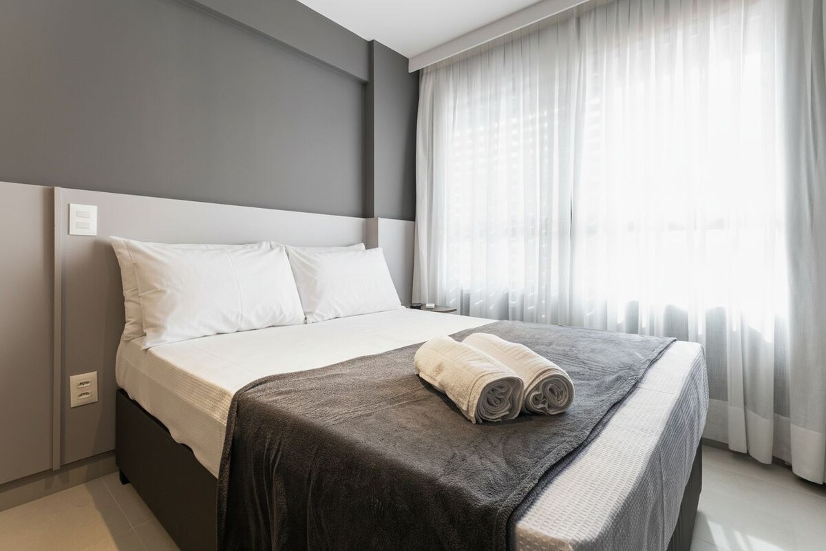 Una habitación de hotel con una cama grande, cabecera, ventana con cortinas y un interruptor de pared.
