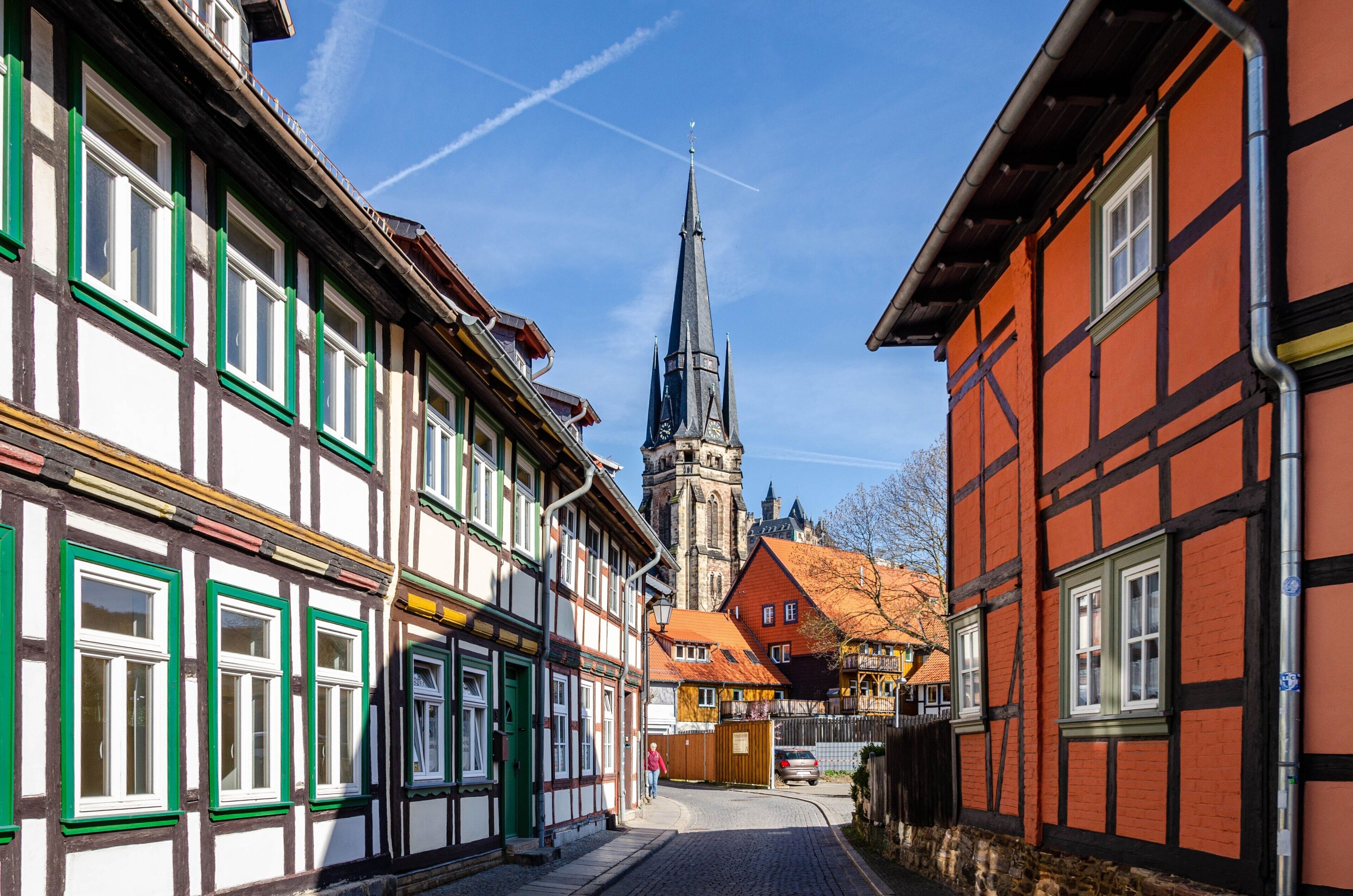 A narrow street lined with traditional half-timbered buildings, a church spire in the distance, and a cobblestone road.