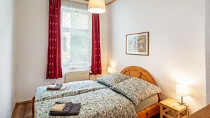 Une chambre avec deux lits, une fenêtre avec des rideaux rouges, une tête de lit en bois, une table de chevet avec une lampe et un tableau encadré au mur.