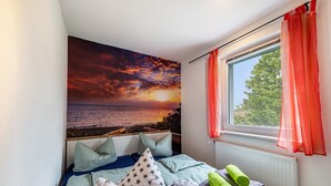 A bedroom with a bed, pillows, a window with curtains, and a wall mural of a sunset over the sea. - Apartment for 6 Guests (57 m²) with Private Terrace and Wi-Fi (Kalkhorst)