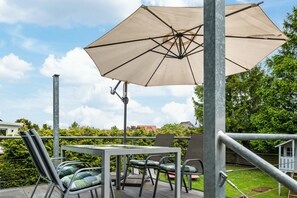 Un patio al aire libre con una mesa y sillas, un paraguas grande, y vistas a un jardín y edificios al fondo.