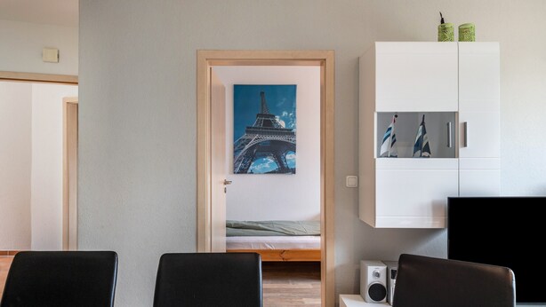 A room with a bed, a dining table with chairs, a white cabinet with pictures, and a painting of the Eiffel Tower. - Apartment for 6 Guests (57 m²) with Private Terrace and Wi-Fi (Kalkhorst)