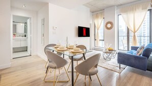 Una sala de estar moderna con una mesa de comedor de vidrio, asientos cómodos, un televisor de pantalla plana y vistas a la ciudad a través de amplias ventanas.