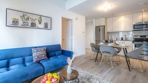 Una sala de estar moderna con un sofá azul, un comedor con sillas grises y una cocina con electrodomésticos de acero inoxidable.
