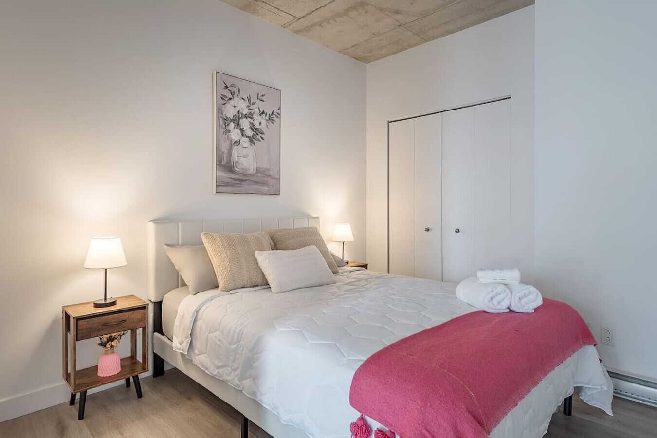 A bedroom with a bed, bedside tables, a nightstand, a painting, a wardrobe, and a red throw blanket.