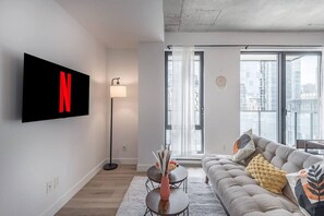 A modern living room with a large TV, a floor lamp, a sofa, and a view of the city through large windows. - Chic Loft Near Old Port | Prime Spot (Montreal)