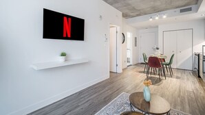 A modern living space with a mounted TV displaying a red 'N', a dining area with a table and chairs, and a small plant on a shelf. - Modern 1BR in Old Montreal + Lounge Access (Montreal)