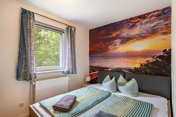 Ein Schlafzimmer mit Bett, einem Fenster mit Vorhängen, einem Heizkörper, einem Nachttisch, einer Lampe und einem großen Gemälde mit Sonnenuntergang an der Wand.