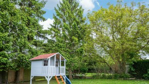 A wooden playhouse with a red roof, a ladder, and a slide, surrounded by a grassy yard and trees.
