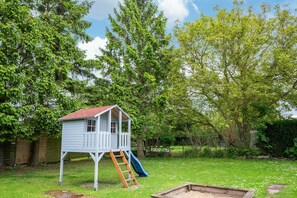 Ein hölzernes Spielhaus mit rotem Dach, einer Leiter und einer Rutsche, umgeben von einem Rasen und Bäumen.