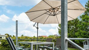 An outdoor patio with a table and chairs, a large umbrella, and a view of a garden and buildings in the background.