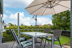 En udendørs terrasse med et bord og stole, en stor parasol samt udsigt til en have og bygninger i baggrunden.