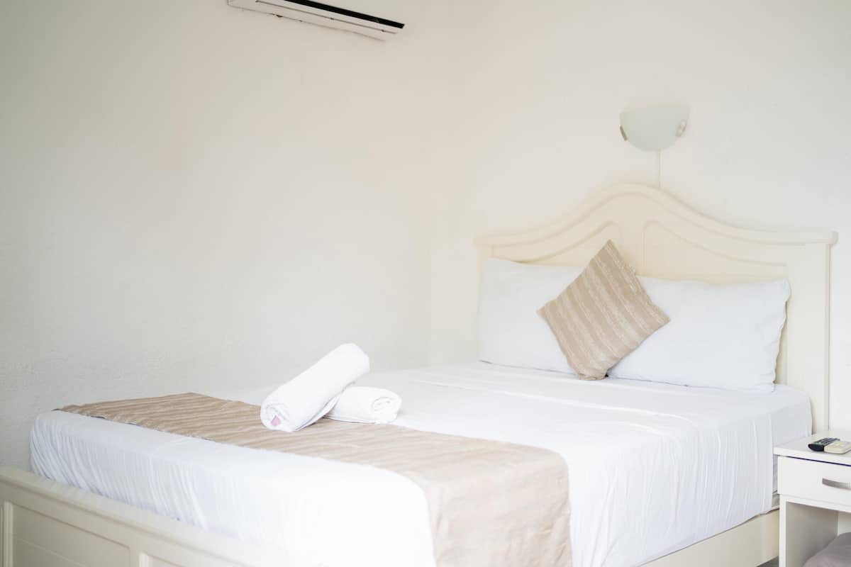 Una cama bien tendida con una colcha blanca y beige, una toalla enrollada al lado y una mesita de noche con una lámpara.