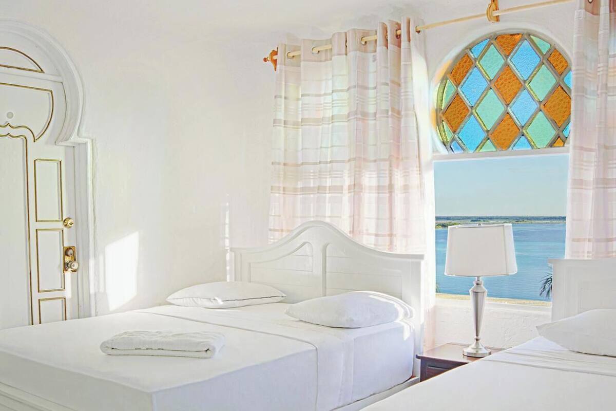 Una cama blanca con cabecera, una ventana con vista al mar y una lámpara sobre una mesita de noche.